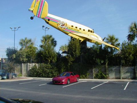 1994 Ford Probe - A red sports car is parked in a lot under a yellow airplane with colorful stripes.