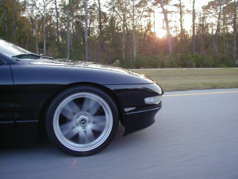 1994 Ford Probe - A black sports car is captured in motion on a highway as the sun sets through the trees.
