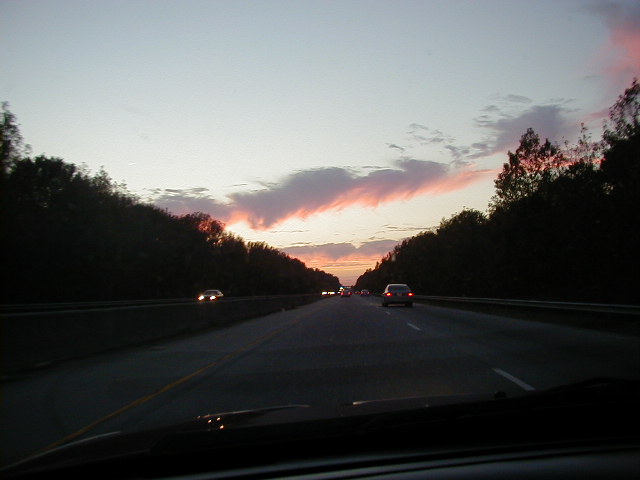 Highway scene at dusk with a pink and purple cloudy sky and trees lining the road