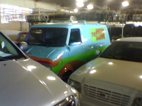 1988 Ford Bronco - The iconic Scooby-Doo Mystery Machine van is seen parked indoors, partially obscured by other vehicles.