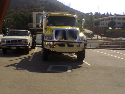 1988 Ford Bronco - Two trucks are parked in a lot, with a building and hills in the background.