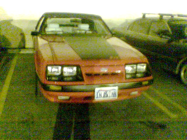 Grainy front view of a red Fox-body Ford Mustang with quad headlights parked in a lot