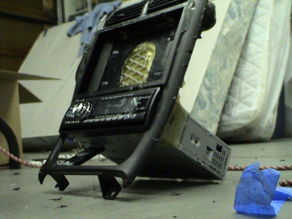 Car stereo and black dashboard trim piece with vents and a gold honeycomb pattern behind them. Some mattresses are in the background
