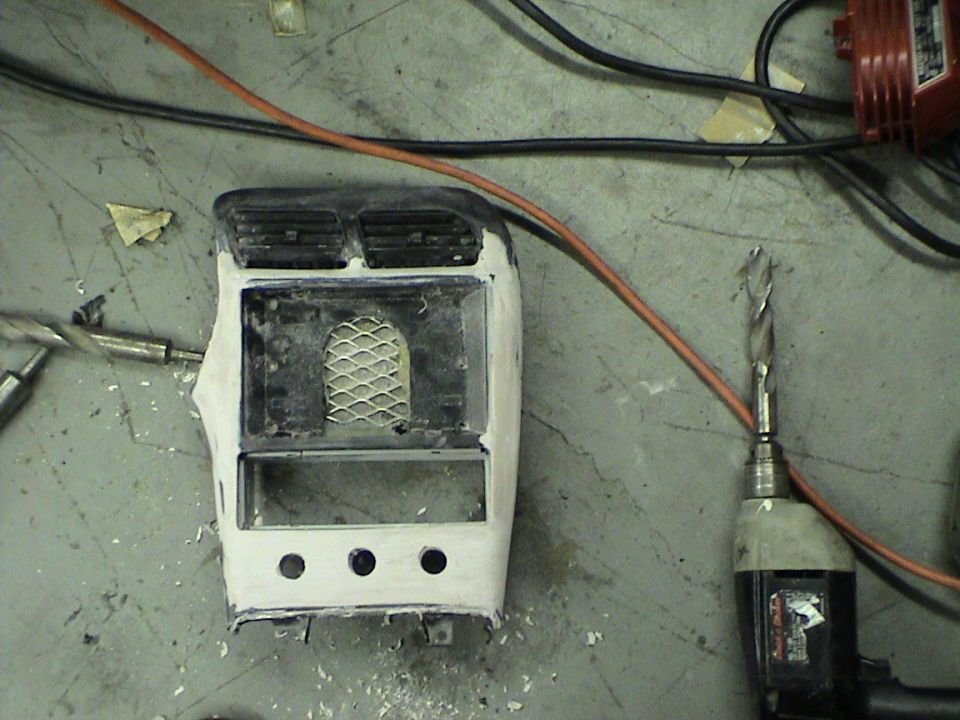 Car dashboard center console piece with air vents, mesh, and three circular cutouts, next to a drill