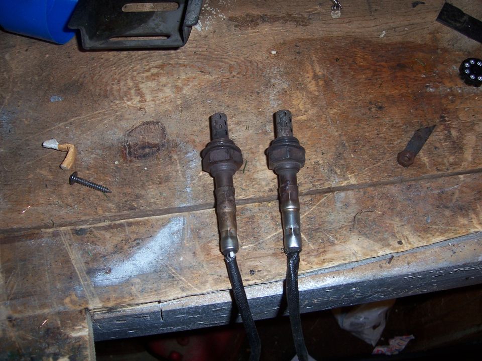 Two used oxygen sensors on a wooden workbench with a screw and other small items