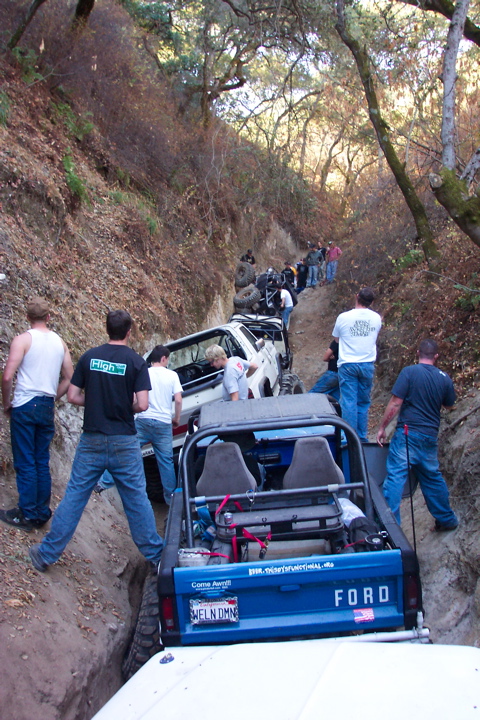1989 Ford Bronco - A line of off-road vehicles navigates a tight trail through a wooded area.