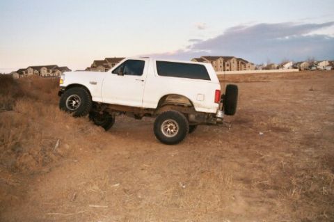 1993 Ford Bronco - This 1993 Ford Bronco is shown off-roading on a dirt hill, with residential buildings visible in the distance.