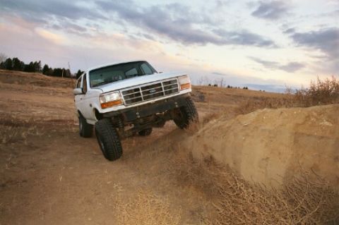 1993 Ford Bronco - A white 1993 Ford Bronco navigates an off-road obstacle under a cloudy sky.