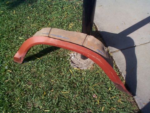 1979 Ford Bronco - A single fender trim piece, painted red, lies on the grass near a sidewalk and a metal pole.