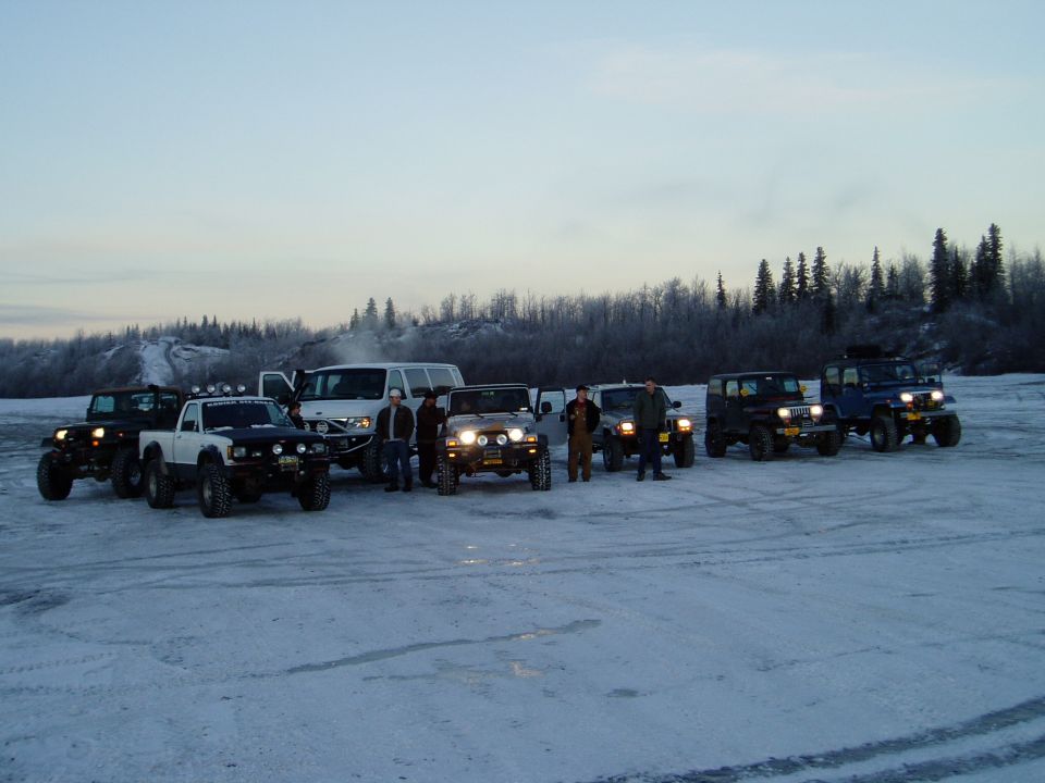 Row of Jeeps and trucks with a white van parked on snow at dusk, people standing between them