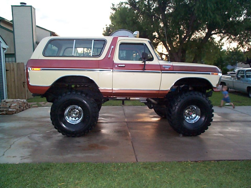Lifted 1978 Ford Bronco with two-tone paint, large off-road tires, and a child running in the background near another pickup truck