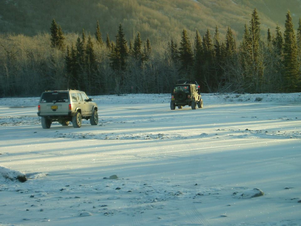 Silver Jeep Cherokee and green Jeep Wrangler driving across a snowy gravel flat near frosted trees