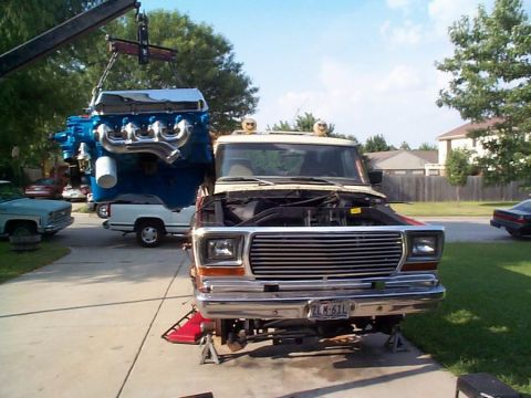 1979 Ford Bronco - A crane is lowering a large blue engine into the engine bay of a truck that is raised on jack stands.