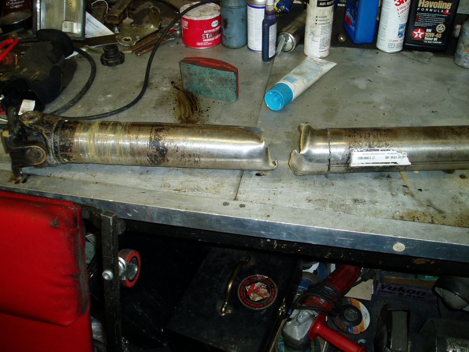 Two driveshafts on a garage workbench with oil cans and chemical bottles behind