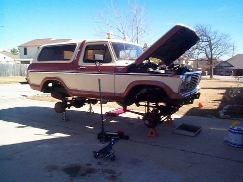 Two-tone brown Ford Bronco on jack stands in a driveway, hood up and wheels off for repair