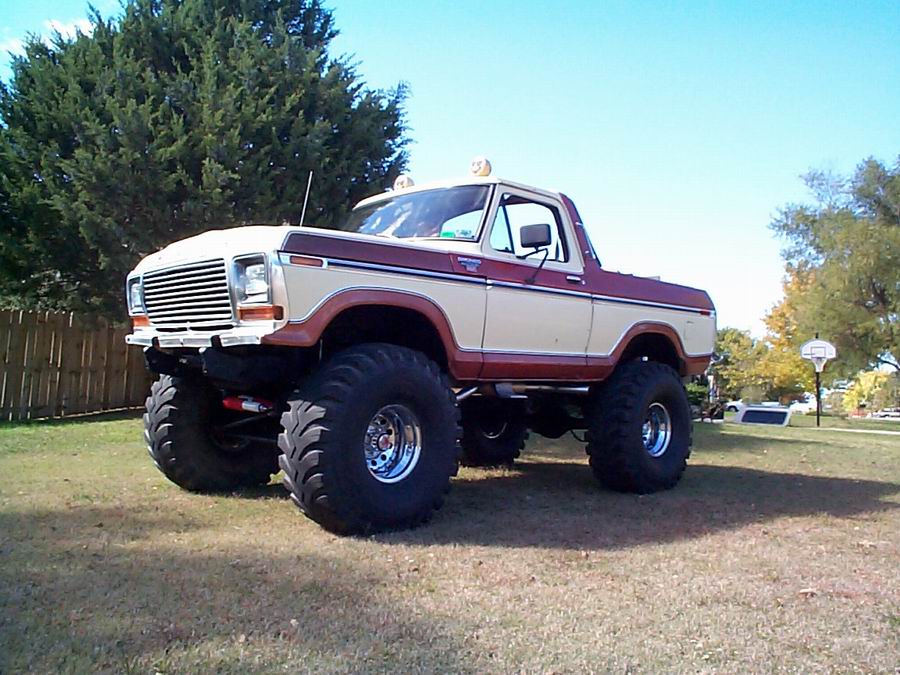 Lifted tan and burgundy Ford Bronco with top removed on big mud tires, 3/4 front view on grass