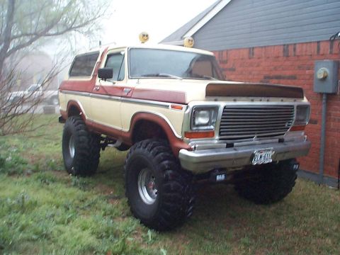 1979 Ford Bronco - A two-tone tan and brown 1979 Ford Bronco full-size SUV lifted on large mud-terrain tires with chrome wheels, parked on grass beside a bric…