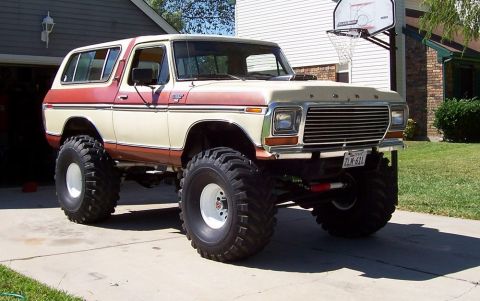 1979 Ford Bronco - A two-tone tan and red-orange 1970s Ford Bronco SUV lifted on tall off-road tires with white wheels, parked on a driveway in a 3/4 front vi…