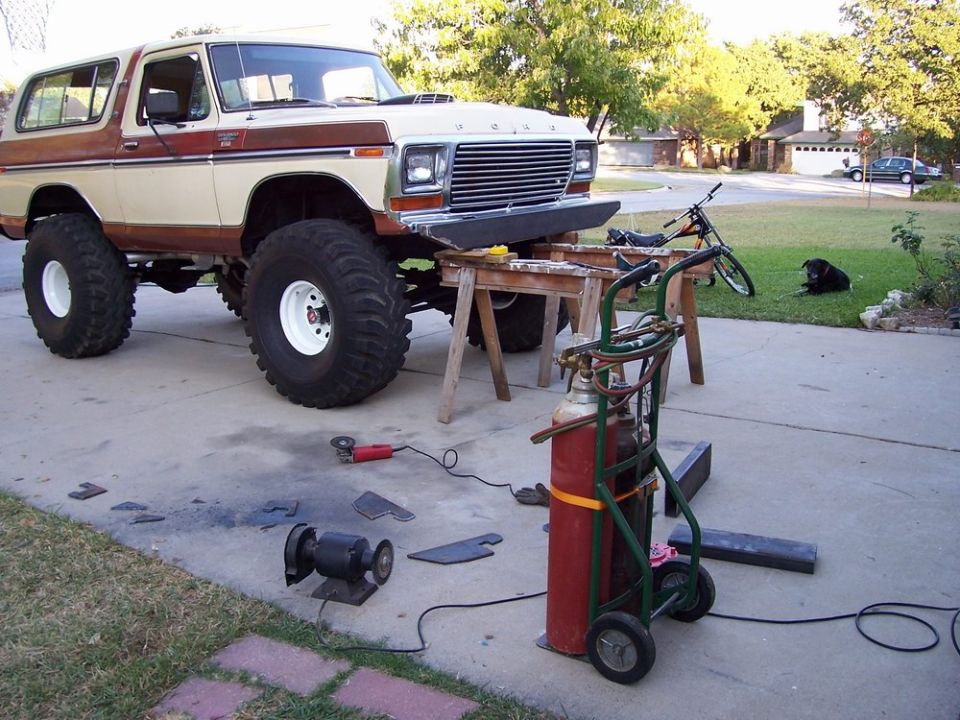 Lifted Ford Bronco on sawhorses, with welding equipment and metal pieces nearby