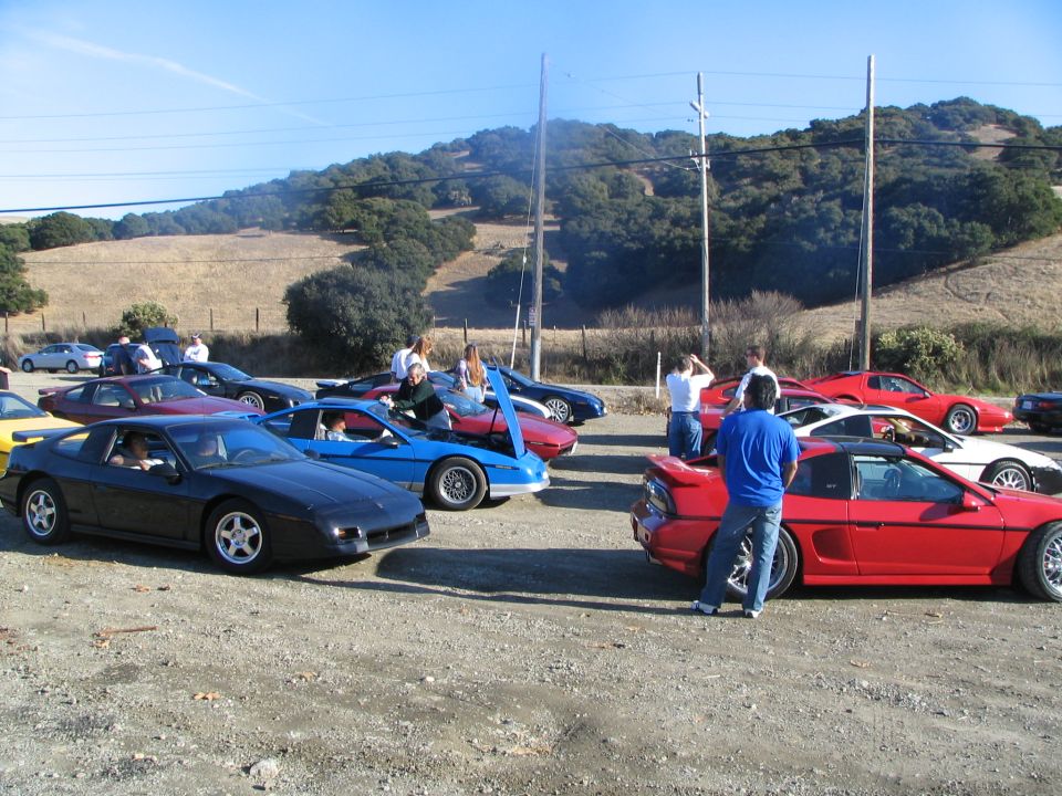 Several Pontiac Fieros parked together, one black, one blue, one red. Other sports cars also present