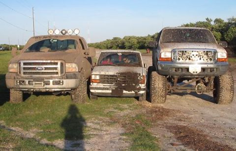 2000 Ford Excursion - Three muddy trucks are parked side-by-side on a grassy area under a clear sky.