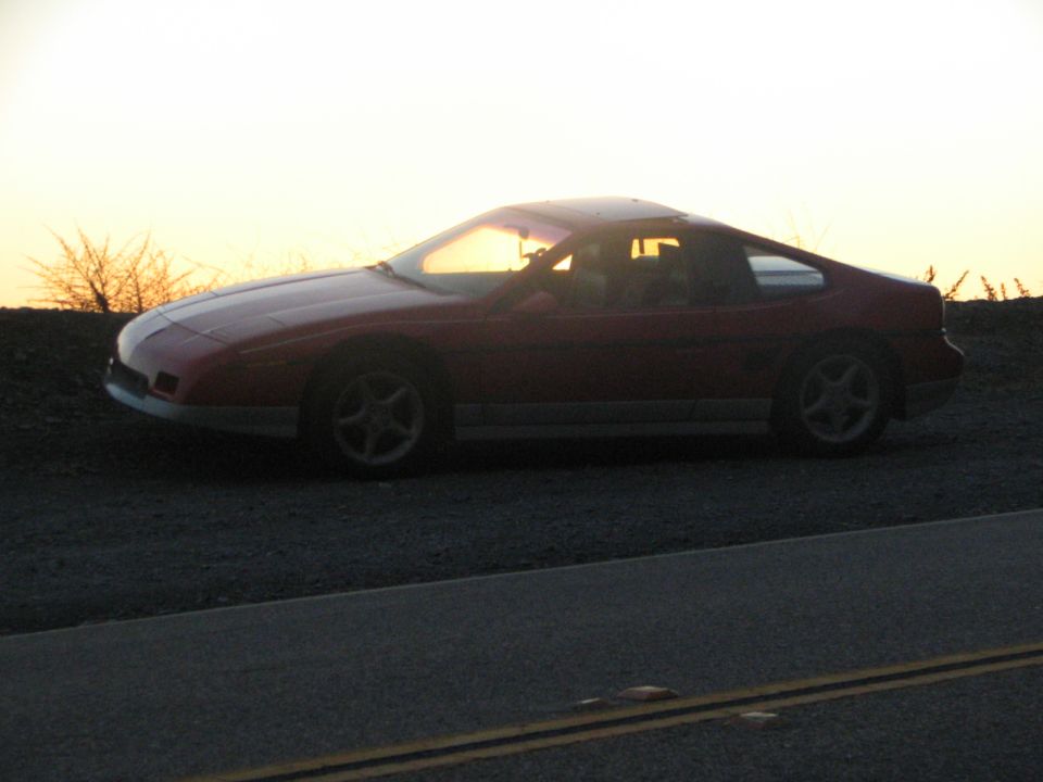 Red 1986 Pontiac Fiero parked on a dirt road at sunset