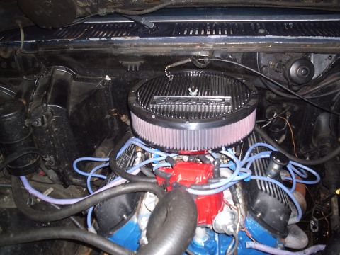 1985 Ford Bronco - Overhead view of a carbureted Ford V8 in an engine bay, topped with a finned chrome air cleaner over a pleated filter element, a red HEI-st…