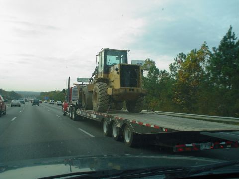 1966 International School Bus - A yellow front loader is transported on a flatbed trailer down a highway, with other vehicles visible in the background.