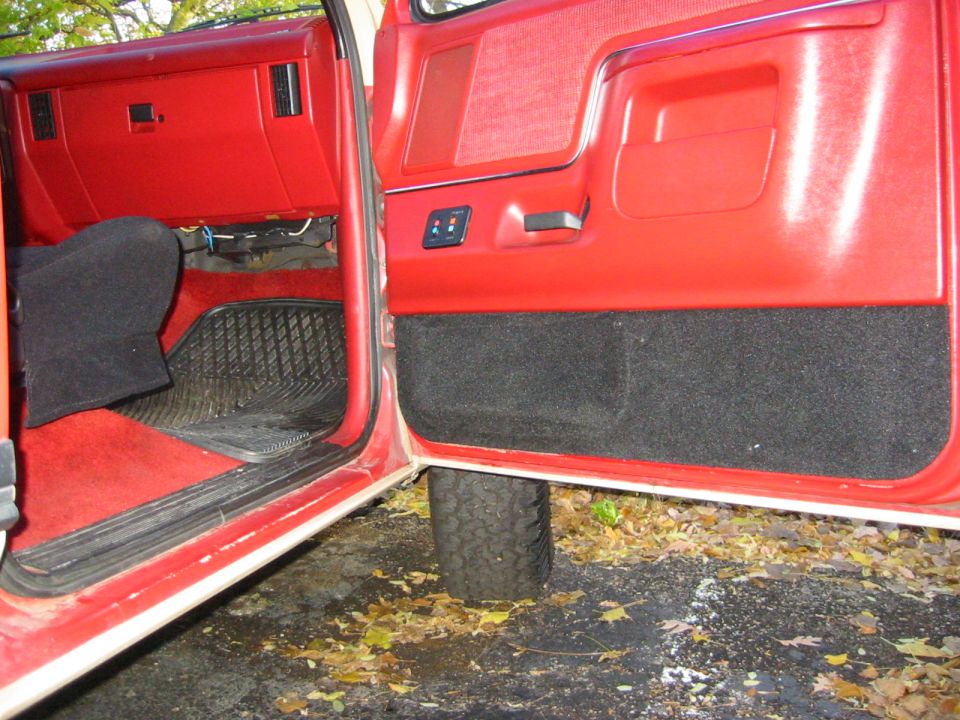 Interior view of a red vehicle, showing the dashboard, door panel, and floor mats. A tire is visible outside the open door