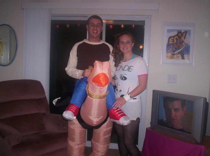 guy in inflatable penis costume with woman next to him, tv shows a man's face