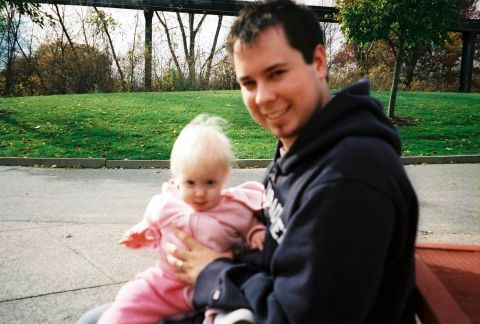 1989 Ford Bronco - A man smiles while holding a baby in a pink outfit, with a park-like setting behind them.