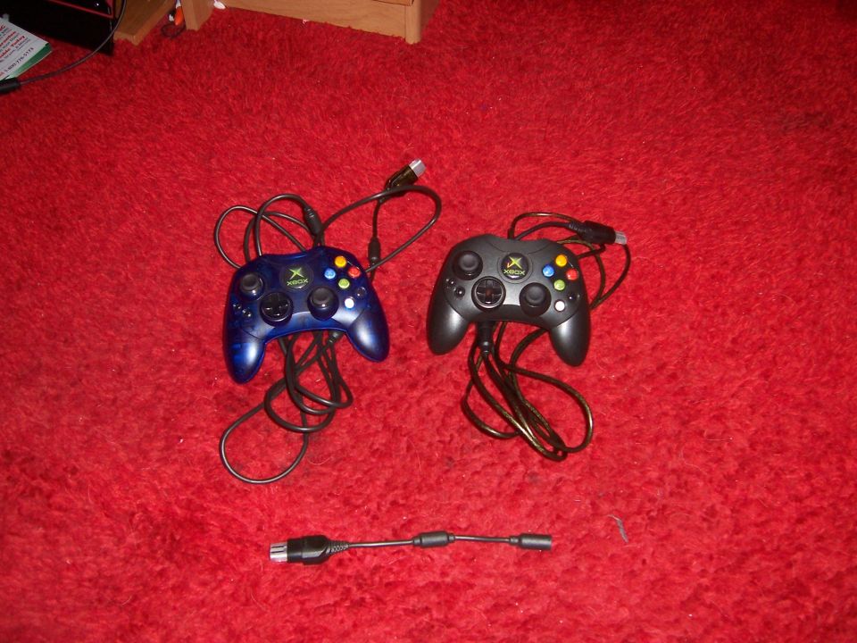 Two wired Xbox controllers, one blue and one black, sit on a red carpet next to an adapter cable