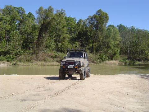 1996 Jeep Cherokee - A tan off-road vehicle with oversized tires sits on a sandy bank next to a body of water.