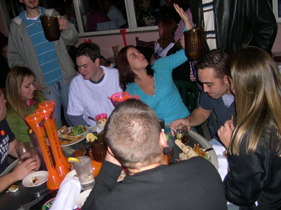 Group of people gathered around a table with large novelty drinks and food, some looking at a woman holding a drink aloft