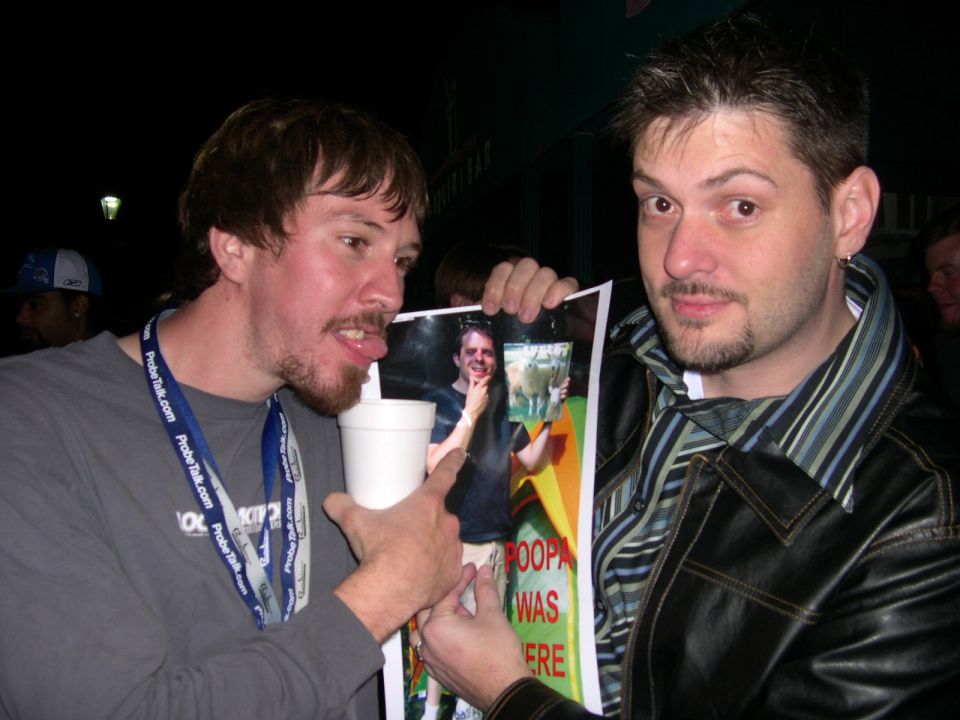Man with tongue out drinks from cup while pointing at a printed photo held by another man