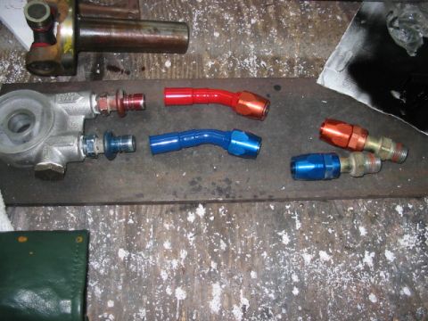 1997 Ford Crown Victoria - Various automotive fluid fittings are laid out on a workbench, including an oil filter housing and several colored AN adapters.