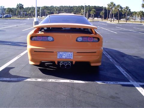 1995 Ford Probe - This orange sports car has a custom body kit and dual exhaust pipes, parked in an empty lot.