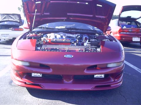 1995 Ford Probe - The front of a red Ford Probe is shown with its hood open, revealing the engine bay. Other vehicles are visible in the background.