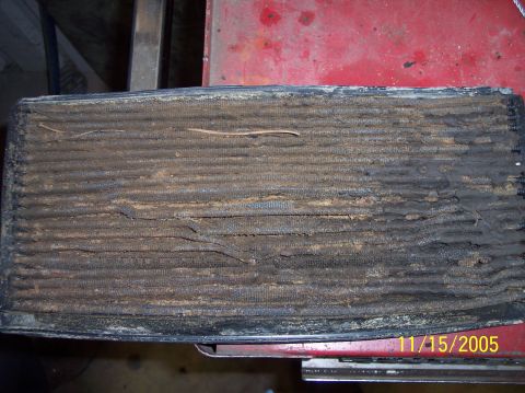 1987 Ford Bronco - A removed radiator core laid on a red tool box, its fins packed solid with caked mud, dirt and bits of pine straw across the full face. A y…