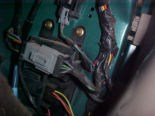 Close-up of a gray electrical connector with multiple wires plugged into it, attached to a green painted surface