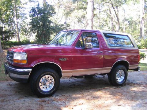 1994 Ford Bronco - This red Ford Bronco is shown from the side, parked outdoors with trees surrounding it.