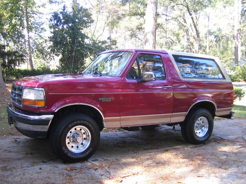 Red Ford Bronco parked on a dirt driveway with trees in the background. It has a tan trim and a removable hardtop
