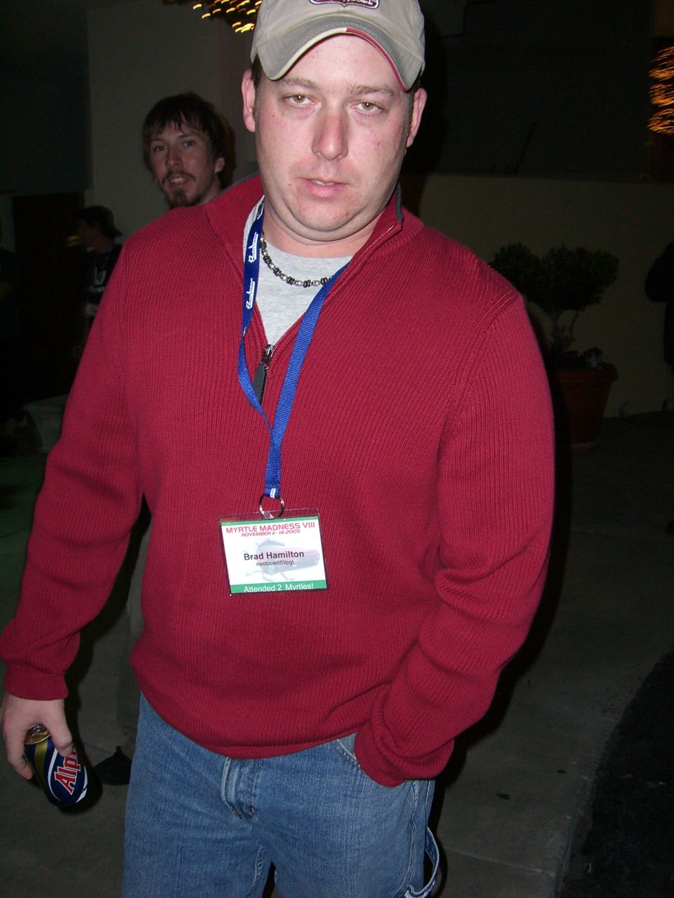 Man wearing a red sweater, blue jeans, and a baseball cap, holding a can of soda. He has a lanyard with a name tag that reads 'Brad Hamilton'