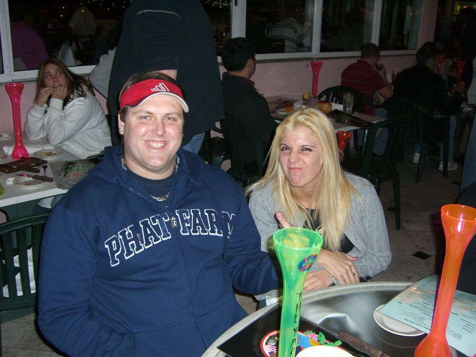 Two people sit at a table in an outdoor restaurant with pink walls and orange and green cups