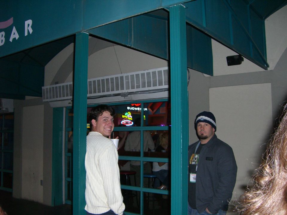 Two men stand outside a bar with a Budweiser sign visible through the window