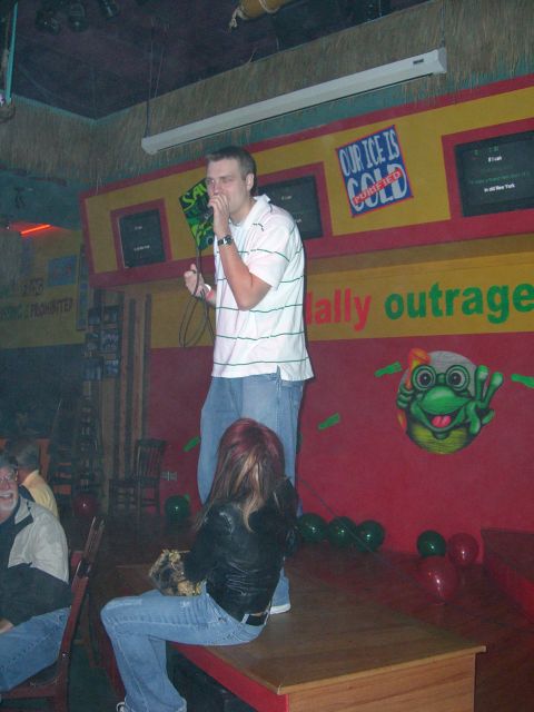 1996 Ford Probe - A man sings into a microphone on a stage, while another person sits nearby. The background features colorful decorations and signs.