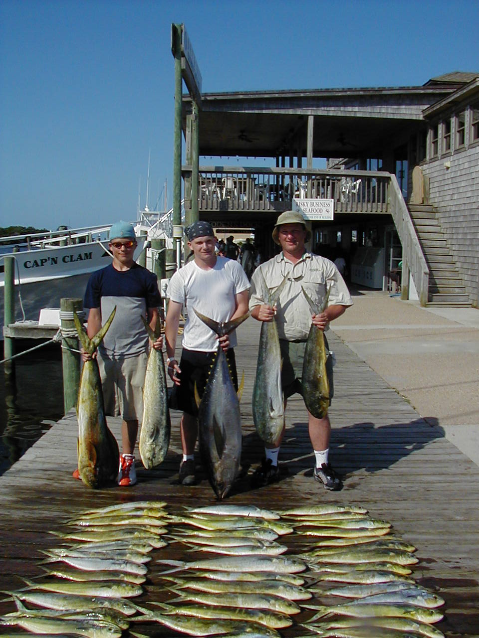 Three men on a dock holding mahi-mahi with rows of fish laid out in front, charter boats behind