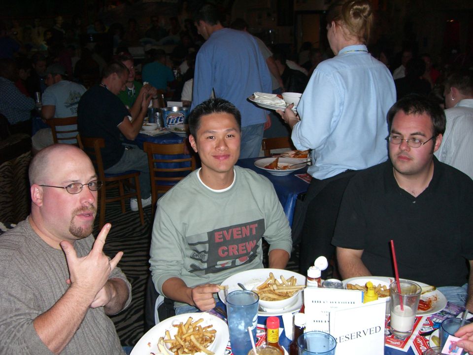 Three men sit at a table with food and drinks, one making a hand gesture