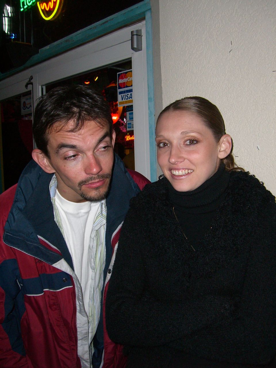 Man in red and blue jacket and woman in black sweater stand in front of a building entrance with credit card logos