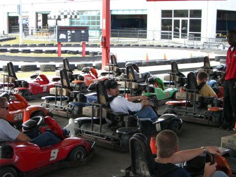1996 Ford Probe - Several go-karts are parked in a line at a track, with people seated in some of them, ready to race.